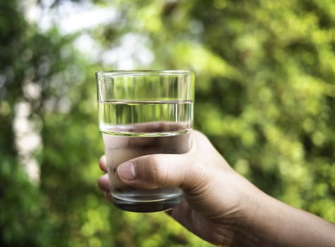 Close-up hand of woman holding cold water in clear glass over nature background.