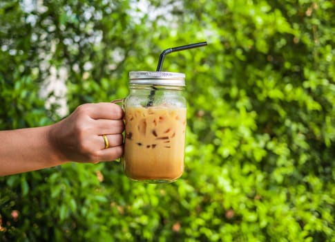 Close-up hand of woman holding iced coffee in clear glass over nature background.
