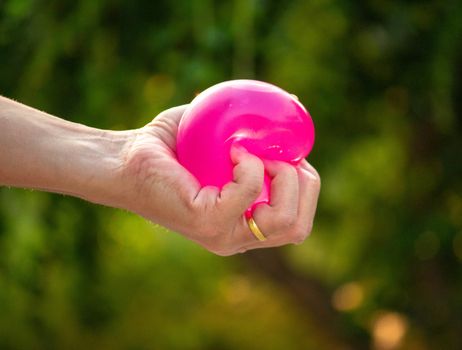 Close-up of hand squeeze the balloons that water inside and splashing around it; on blurred nature background in the garden.