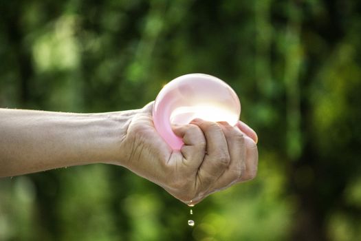 Close-up of hand squeeze the balloons that water inside and splashing around it; on blurred nature background in the garden.