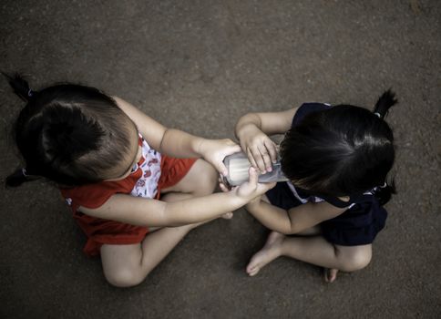 Asian little child sit in the garden and feed some milk to her sister from glass