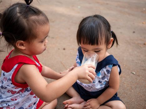 Asian little child sit in the garden and feed some milk to her sister from glass