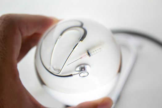 Close-up of Hand holding Crystal glass ball sphere revealing the inner medical stethoscope and syringe on grey gradient background.