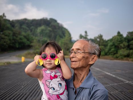 Asian grandfather carrying grandchild while her wearing pink glasses and smile.
