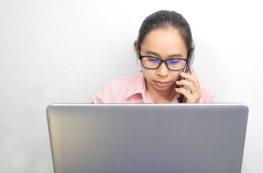 Business Asian young woman talking on the mobile phone while sitting at her working place in office and looking at laptop.