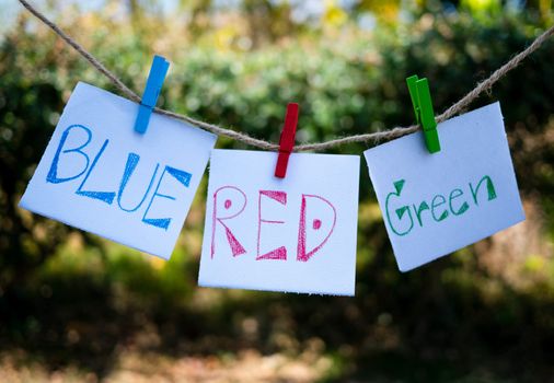 Paper with handwriting word colors, Blue, Red, Green hanging on a  rope with wooden clip and developed in the wind on green nature background.
