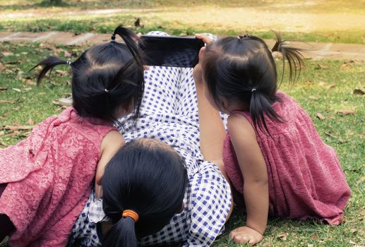 Asian mother and her daughters sitting on the grasses ground in the garden and looking at smartphone happily.