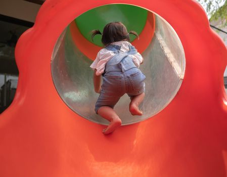 Asian little girl Tried to climb into the red tunnel tube slide at park playground. Playing is learning for children. Dangerous in children concept.