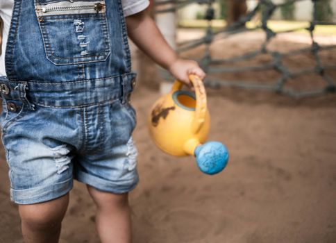 Close up of Asian little girl holding plastic watering can toys in the playground and play a sand with plastic beach toys happily. Playing is learning for children.