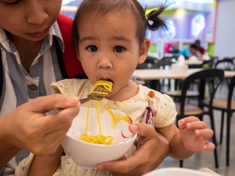 Asian mom feeding the noodle for the daughter to eat with a fork at food court in the mall.