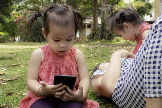Asian little girl sitting on the grasses ground in the garden and looking at smartphone happily while mother and sister lying down to play  near.