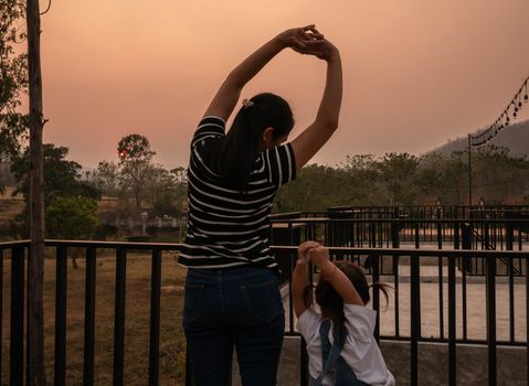 Asian young woman stretch oneself on terrace and looking view sunset in evening with daughter.