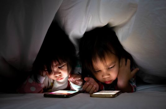 Portrait of Asian little girl with sister look cartoon on a smartphone under a blanket on the bed. The child's face is illuminated by a bright monitor. Health care concept.
