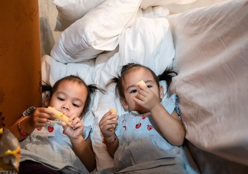 Asian little child girl with sister eating snack deliciously together in thr bed room.