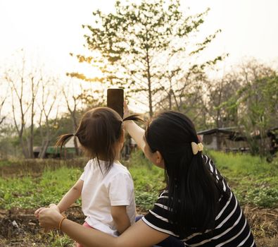 Back view of Asian little child girl taking a photo with mother in garden over sunset background.