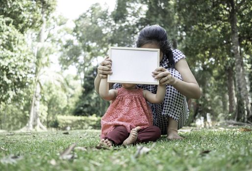 Asian little girl holding empty white board with mom and sitting on grasses ground. Space for write your text.
