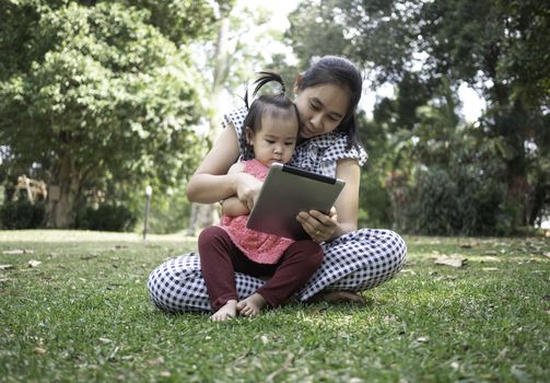 Asian mother and her daughters sitting on the grasses ground in the garden and looking at digital tablet happily.