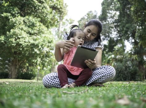 Asian mother and her daughter sitting on the grasses ground in the garden and looking at smartphone happily.