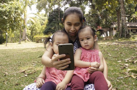 Asian mother and her daughters sitting on the grasses ground in the garden and looking at smartphone happily.