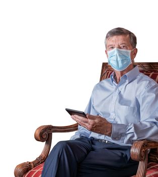 Senior caucasian adult reading from ebook in hand while seated. He is wearing a face mask against coronavirus and cutout against white