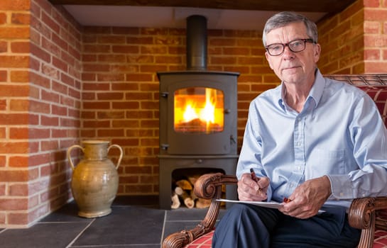 Senior caucasian adult checking a document with pen in hand while seated. He is wearing a pair of glasses and has wood burning fireplace in background