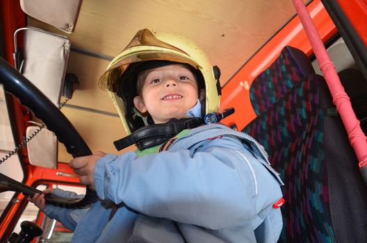 Little boy acting like a fireman. Boy sitting in a real fireman car. Happy adorable child boy with fireman hat sitting in red fire truck looking out. Dreaming of future profession. Fire safety, life protection lessons.