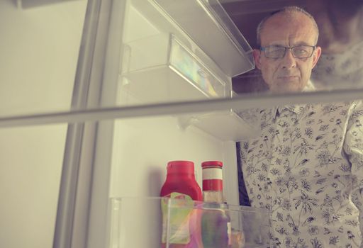 Portrait of a hungry senior man looking for food in refrigerator. Eating and diet concept - confused mature-aged man looking for food in empty fridge at kitchen.