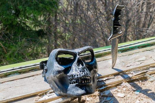 a terrible mask in the form of a skull and an old combat knife stuck in the destroyed windowsill in the window of an abandoned house against the green leaves of a coniferous tree