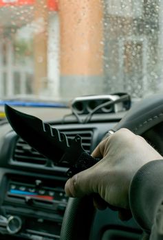 a knife on the steering wheel of a car in the hand of a man against the door of a house and rain drops on the windshield