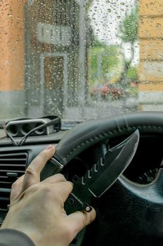 knife on the steering wheel of the car in the hand of a man on the background of the arch of a house and rain drops on the windshield
