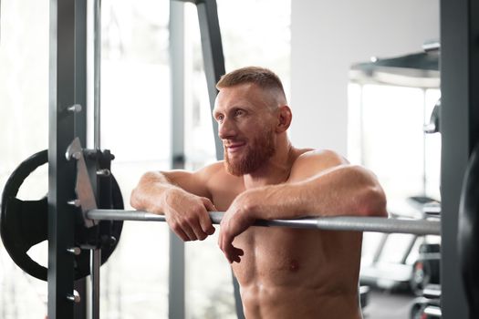 Gym man leaning barbell resting after hard training. Handsome caucasian athlete take break. Sport equipment Sportive guy standing near barbell machine. Perfect muscule male body ideal. Naked torso