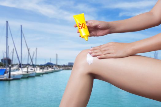 woman applying sunscreen on her leg with blur boats docked at the yacht club background. SPF sunblock protection concept. Travel vacation