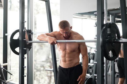 Gym man leaning barbell resting after hard training. Handsome caucasian athlete take break. Sport equipment Sportive guy standing near barbell machine. Perfect muscule male body ideal abs. Naked torso