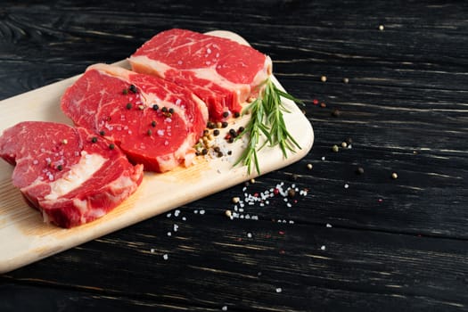 Three pieces of juicy raw beef with rosemary on a cutting board on a black wooden table background. Meat for barbecue or grill sprinkled with pepper and salt seasoning