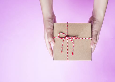 Female hands holding a simple craft paper gift box wrapped with colorful rope with as a present for Christmas, new year, valentine day or anniversary on pink background, top view