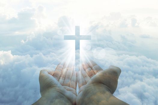 hands cupped with a shining white cross hovering above on a sunny blue daytime sky with fluffy clouds,double exposure image.