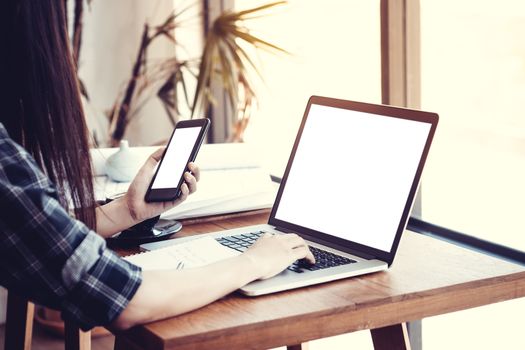 Young Asian woman freelance using smart phone and notebook computer working at home.Girl browsing internet, chatting, blogging. Female holding phone and looking on blank white screen, anonymous face.