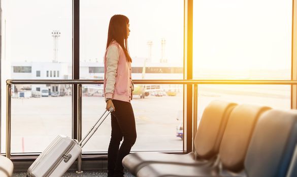 Travel lifestyle concept. Travel tourist woman waiting at boarding gate terminal before departure with luggage looking at airplanes with sunrise view at airport window for morning flight.