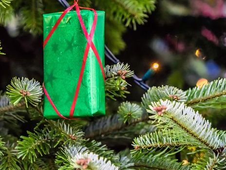 Green wrapped Christmas present with a red ribbon hanging on a Christmas tree, closeup