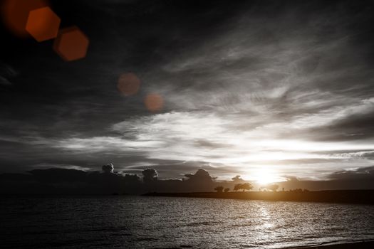 sunset on cloudy sky at the sea beach with silhouette rock bridge and tree background , black and white color with red lens flare effect