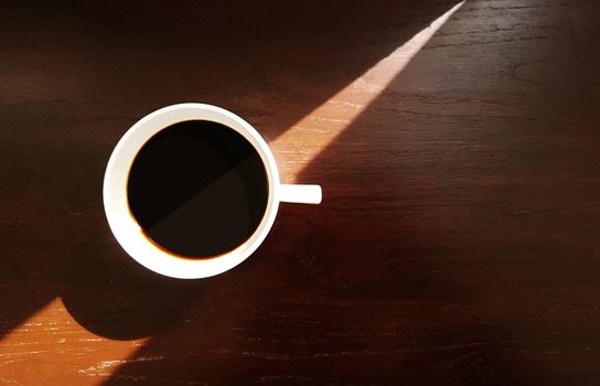 White ceramic coffee cup with espresso black coffee on wooden table background with triangle sun light and shade from window, top view