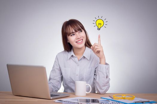 Success growing business concept : Thinking business woman looking up with light idea bulb above finger isolated on gray wall background. Portrait of young Asian model in her 20s