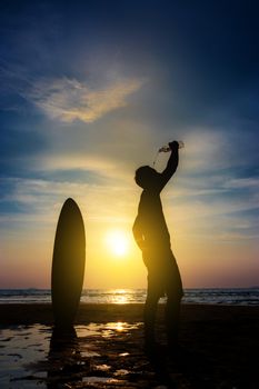 Silhouette of surf man stand with a surfboard drink water from bottle. Surfing at sunset beach. Outdoor water sport adventure lifestyle.Summer activity. Asia male model in his 20s