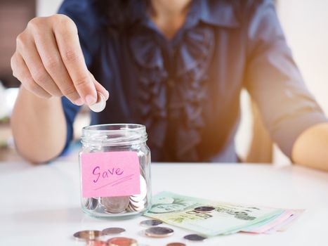 Asian women use hand to collect coins in glass bottles with Thai money banknote resting on white table. Save money concept.