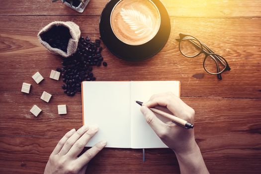 Education background concept : close up man with pencil in hand writing in notebook on wood table with coffee cup, top view. Vintage light effect.