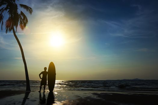 Silhouette of surf man stand with a surfboard and coconut palm. Surfing at sunset beach. Outdoor water sport adventure lifestyle.Summer activity. Handsome Asia male model in his 20s.
