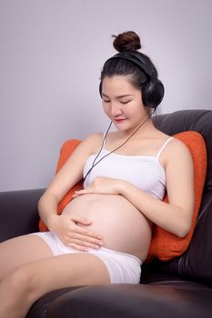 Portrait of pregnant Asian woman sitting on sofa at home and listening music in headphones. Beautiful Asia female model in her 20s.