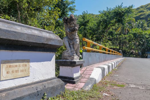 Bridge with yellow metal railing over river in mountains on Bali island. Decorative stone statues represent mythological protectors. In the background wild tropical vegetation and mountain range.