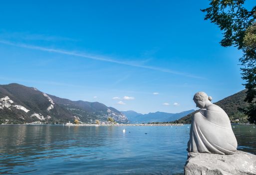 Sculpture of a young woman sitting on a rock, on the shore of Lake Iseo. In the background, the lake and the mountains.