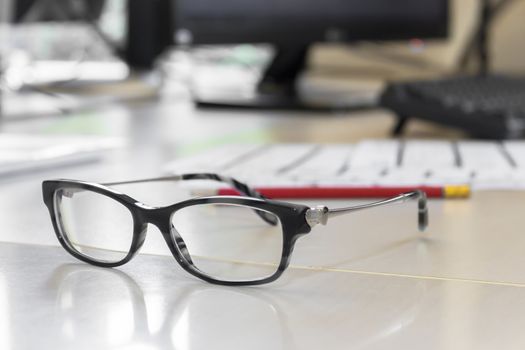 Black eyeglasses on wooden table. On the blurred background they can be seen a pencil and a Desktop pc. Ideal for Business and finance concept of office working.